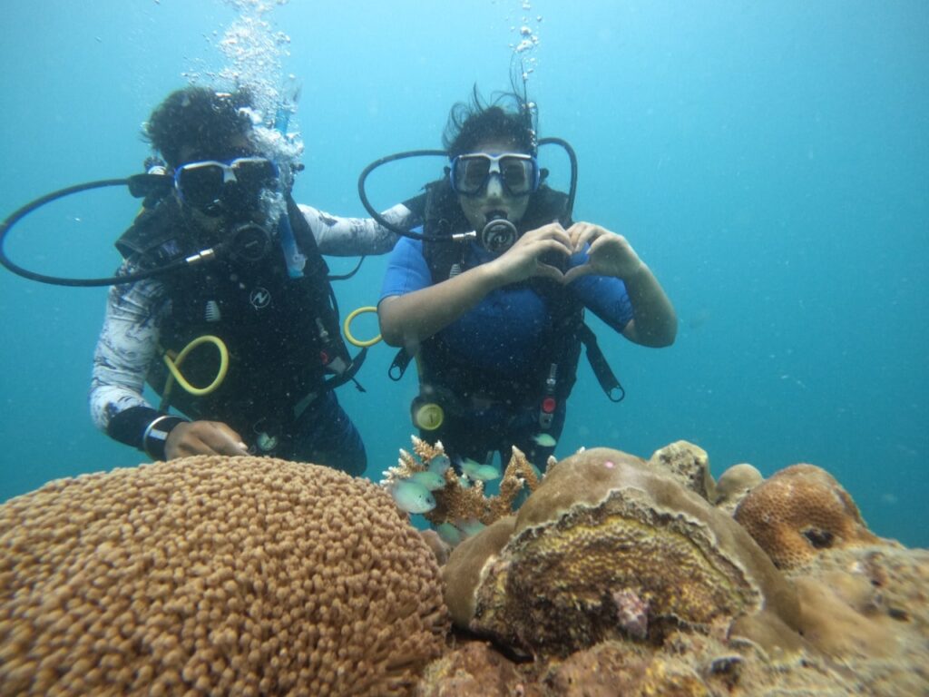 One male and one female scuba divers diving underwater  and in front of them are coral reefs while scuba diving at Tribe gate dive site.  